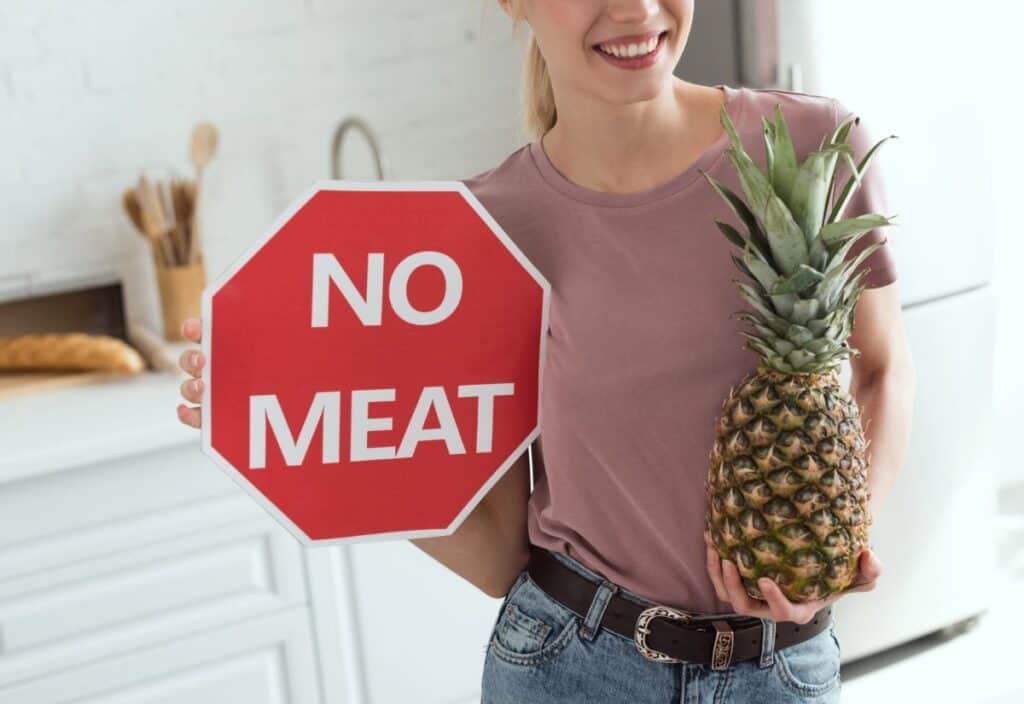 A person holds a "NO MEAT" stop sign and a pineapple while standing in a kitchen.