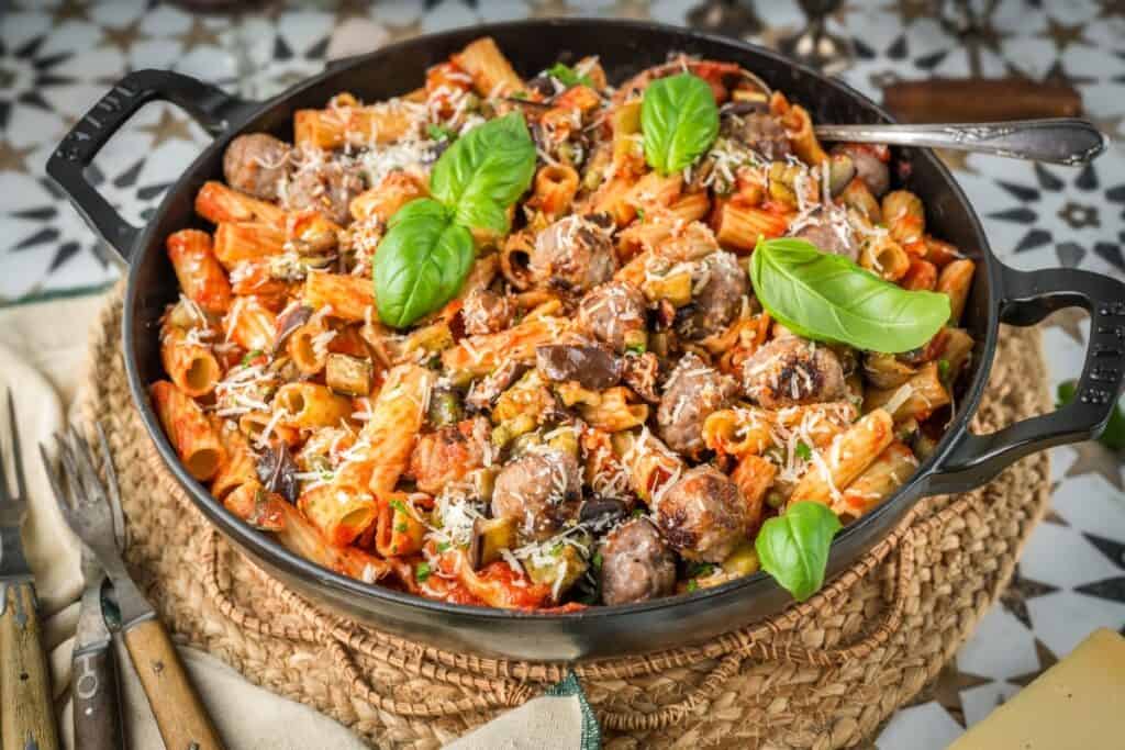 Large pan of baked rigatoni pasta dinner with sausage, tomato sauce, mushrooms, and grated cheese, garnished with fresh basil leaves, on a woven placemat with cutlery beside it.