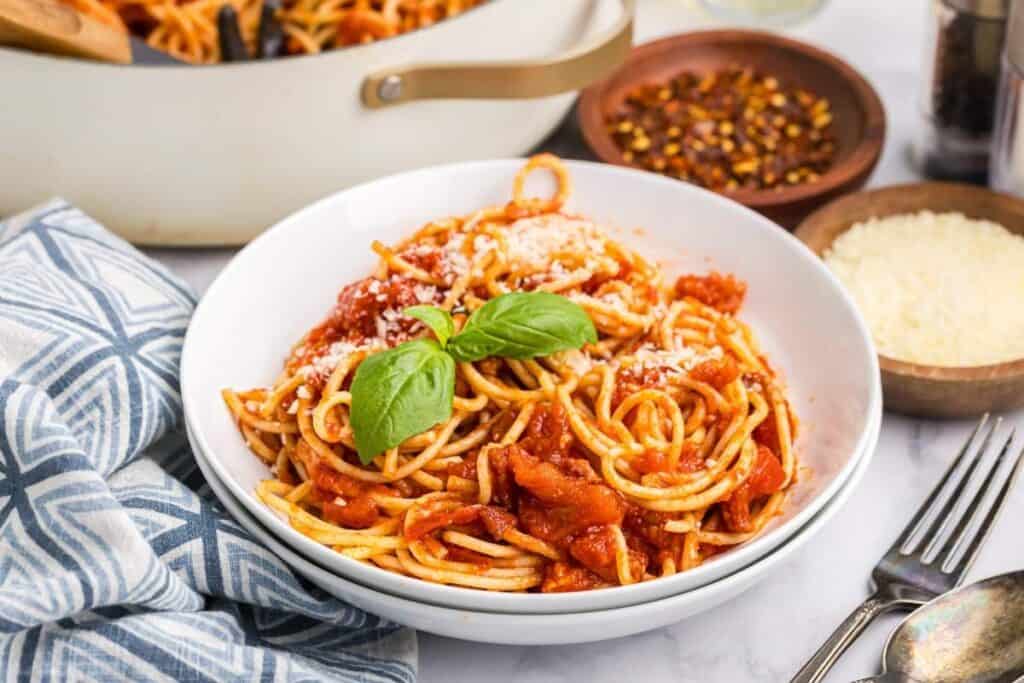 A bowl of spaghetti with tomato sauce, grated cheese, and basil garnish sits on a marble surface next to a napkin and bowls of cheese and chili flakes, creating the perfect pasta dinner setting.