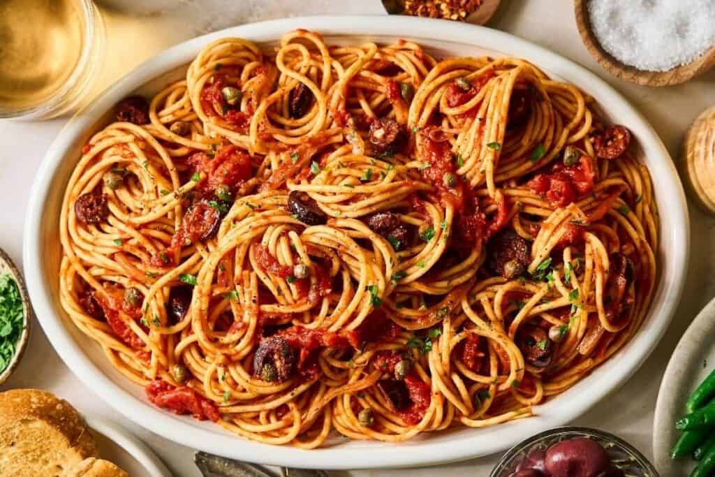 A white oval dish filled with spaghetti tossed in tomato sauce with black olives and chopped herbs, showcasing one of the Pasta Classics.