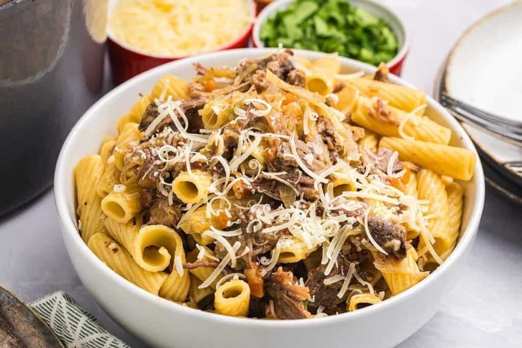 A bowl of rigatoni pasta, one of the Pasta Classics, topped with shredded meat and grated cheese, with bowls of extra shredded cheese and chopped greens in the background.