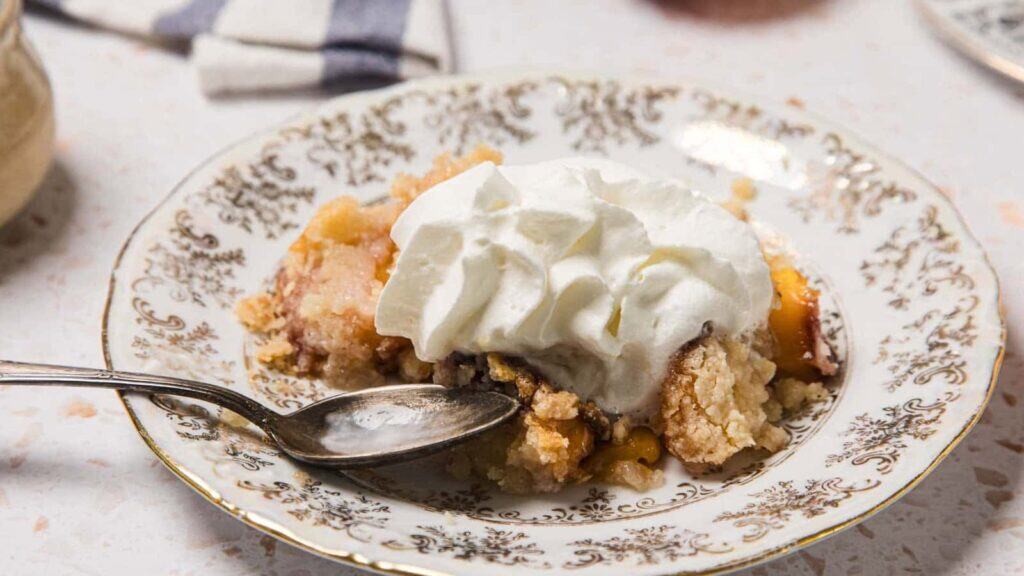 A serving of fruit cobbler topped with whipped cream on a decorative plate, with a spoon resting on the side.
