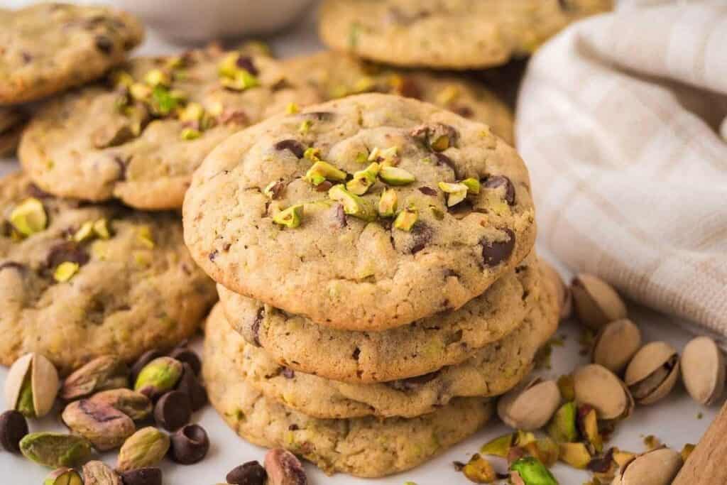 A stack of tasty chocolate chip cookies topped with chopped pistachios, surrounded by more cookies and scattered whole and shelled pistachios.