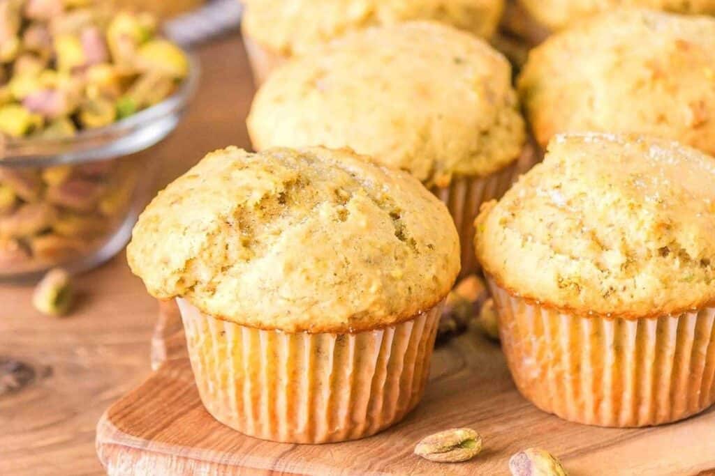 A close-up of several freshly baked muffins, classic baked goods, on a wooden board, with a bowl of chopped pistachios in the background.