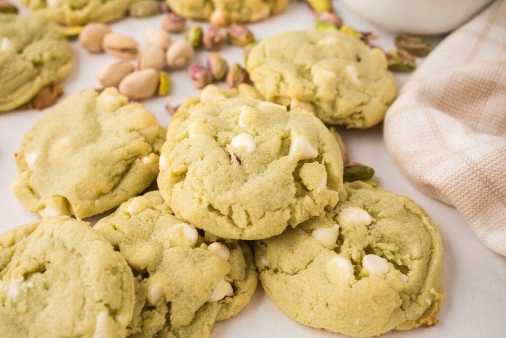 A pile of tasty cookies, filled with pistachio and white chocolate chips, sits on a white surface, surrounded by scattered pistachio nuts and a beige cloth.