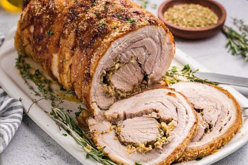 A sliced rolled roast pork with herb stuffing on a white platter, garnished with fresh herbs, with a bowl of seasoning in the background&mdash;perfect for your next Pork Dinner ideas.