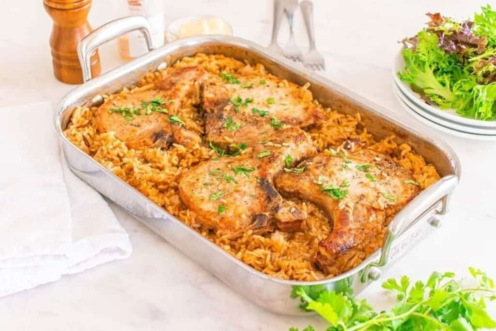 A rectangular baking dish with seasoned pork chops on a bed of cooked rice, garnished with herbs, showcases one of the classic casserole recipes. It rests on a white surface next to a plate of salad and utensils.
