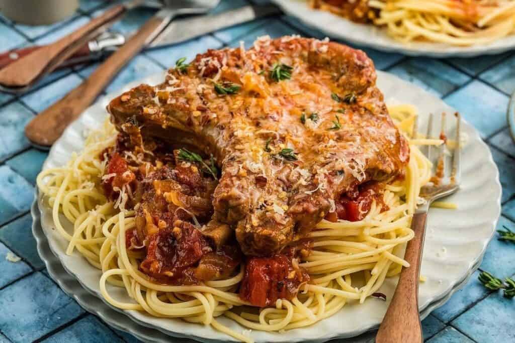 A plate of spaghetti topped with tomato sauce and a seasoned pork chop, garnished with grated cheese and herbs, offers inspiration for delicious pork dinner ideas, served on a table with utensils.