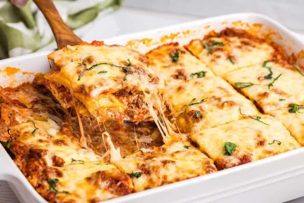 A baking dish of cheesy lasagna, a true Pasta Classics favorite, is being served with a wooden spatula; the lasagna is cut into square portions and topped with melted cheese and herbs.