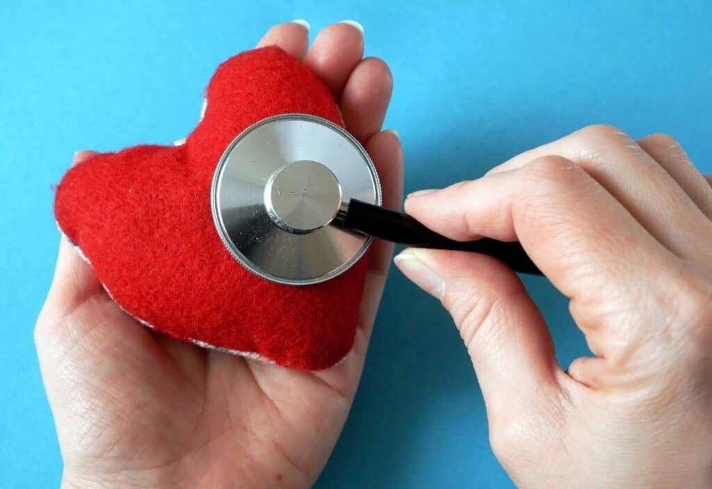 A hand holds a red plush heart while another places a stethoscope on it, symbolizing care and awareness for American Heart Month, set against a blue background.