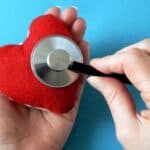 A hand holds a red plush heart while another places a stethoscope on it, symbolizing care and awareness for American Heart Month, set against a blue background.
