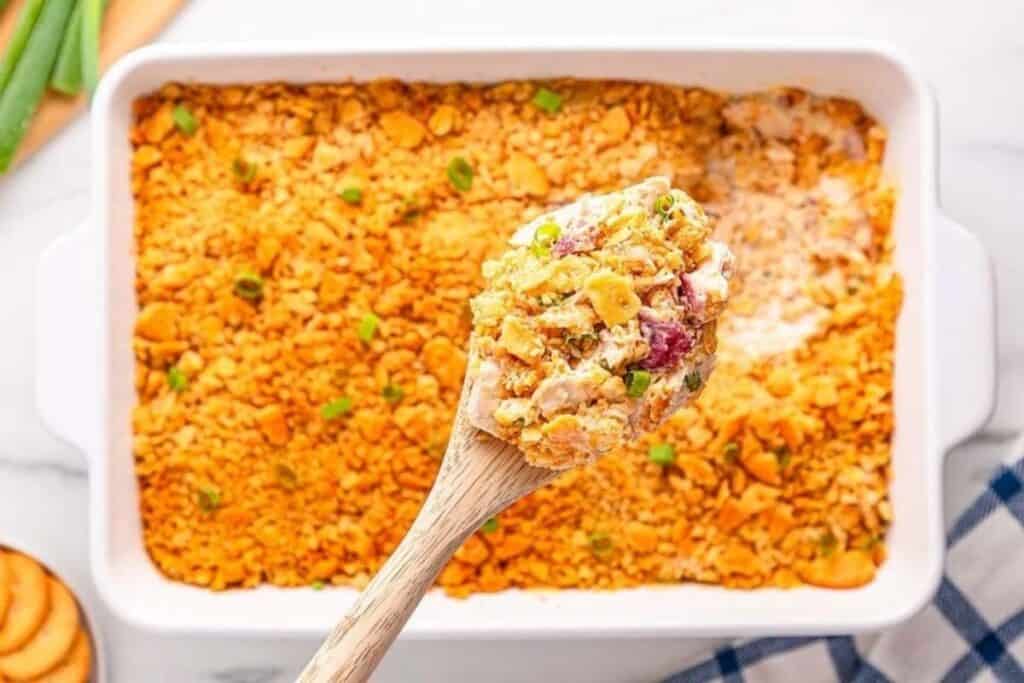 A close-up of a wooden spoon holding a portion of a creamy casserole topped with crushed crackers, shown above the casserole dish&mdash;a delicious highlight from classic casserole recipes.
