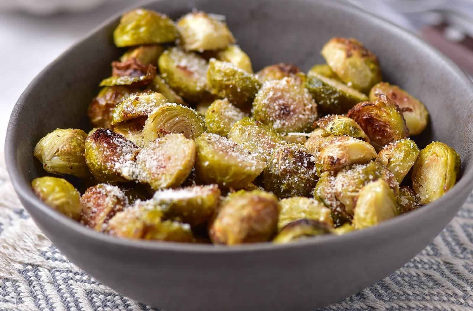 A gray bowl filled with roasted Brussels sprouts sprinkled with grated cheese sits on a patterned tablecloth.