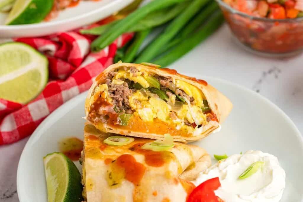 A breakfast burrito cut in half on a plate, filled with eggs, beef, cheese, and vegetables&mdash;served with sour cream, lime wedges, and salsa. One of the tastiest breakfast ideas for a flavorful morning!.