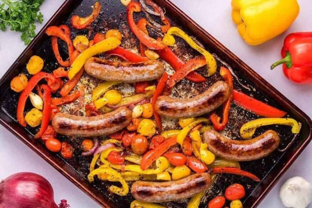 A sheet pan with roasted sausages, cherry tomatoes, and sliced red and yellow bell peppers&mdash;an easy, vibrant option for weeknight dinners. Fresh vegetables are scattered around the pan.