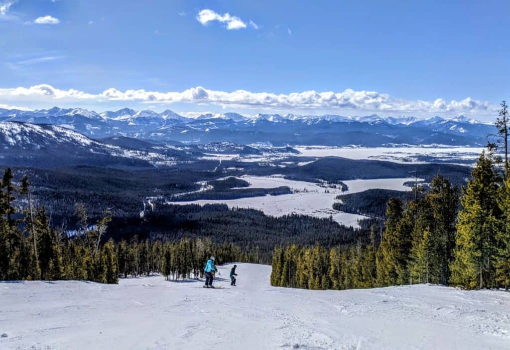Two skiers descend a snowy slope surrounded by pine trees, with a vast mountain range and a valley visible under a clear blue sky in the background.