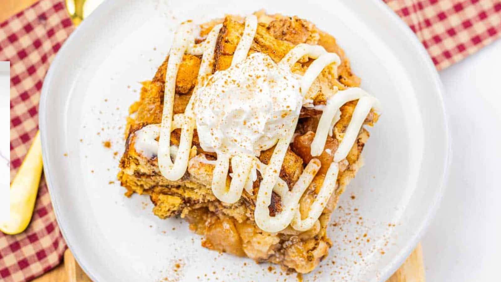 A slice of apple pie with whipped cream on top, served on a white plate with apples in the background.