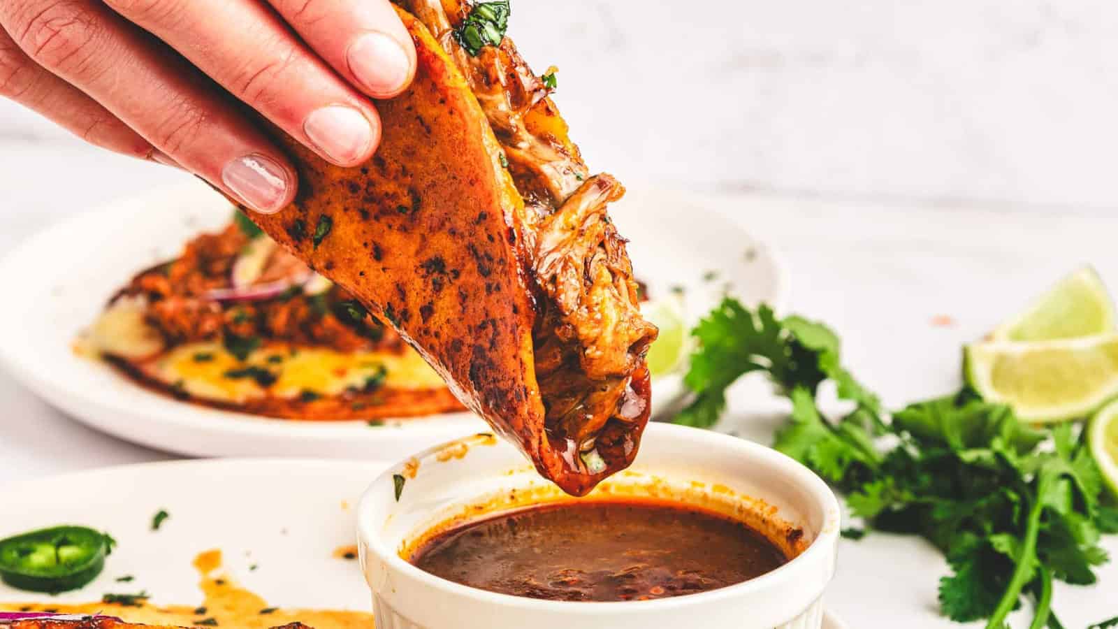 Three birria tacos with lime wedges, sliced onions, and dipping sauce on a white plate.