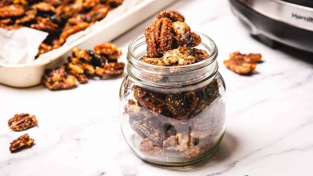 A glass jar filled with candied pecans sits on a white surface, with more candied pecans scattered nearby and a tray of them in the background.