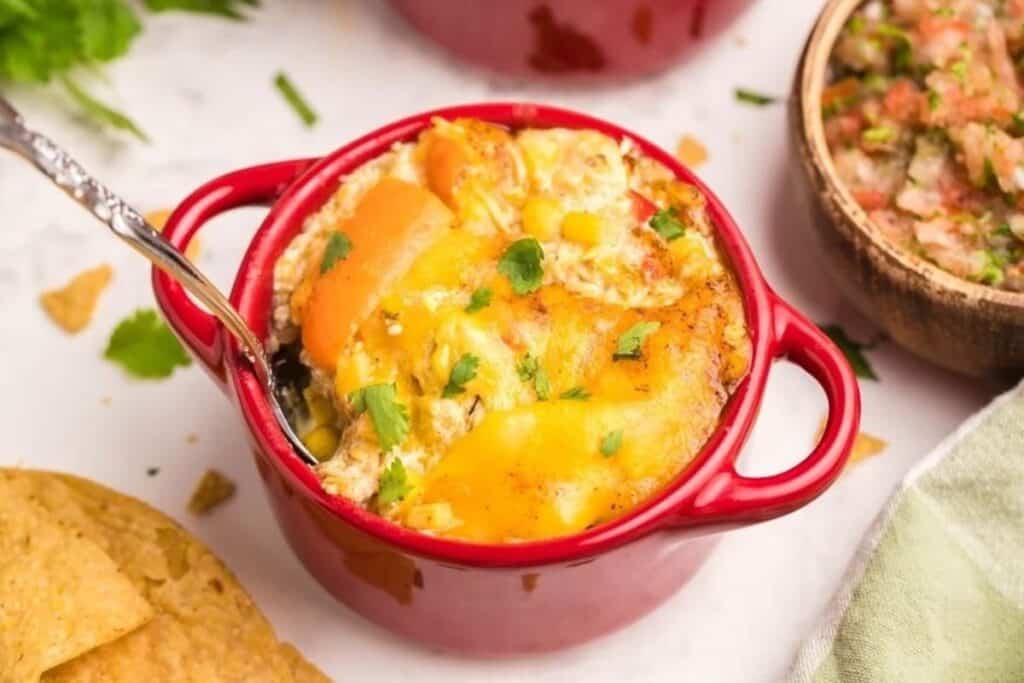 A small red dish filled with cheesy casserole, inspired by classic casserole recipes, is garnished with cilantro and sits next to tortilla chips and a bowl of salsa.