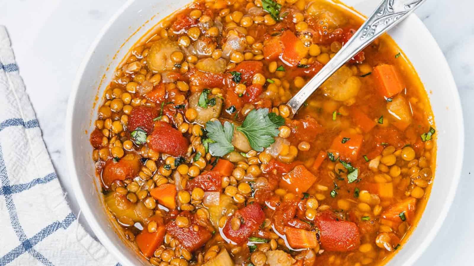 A bowl of lentil soup with carrots, tomatoes, herbs, and a spoon, garnished with fresh parsley.