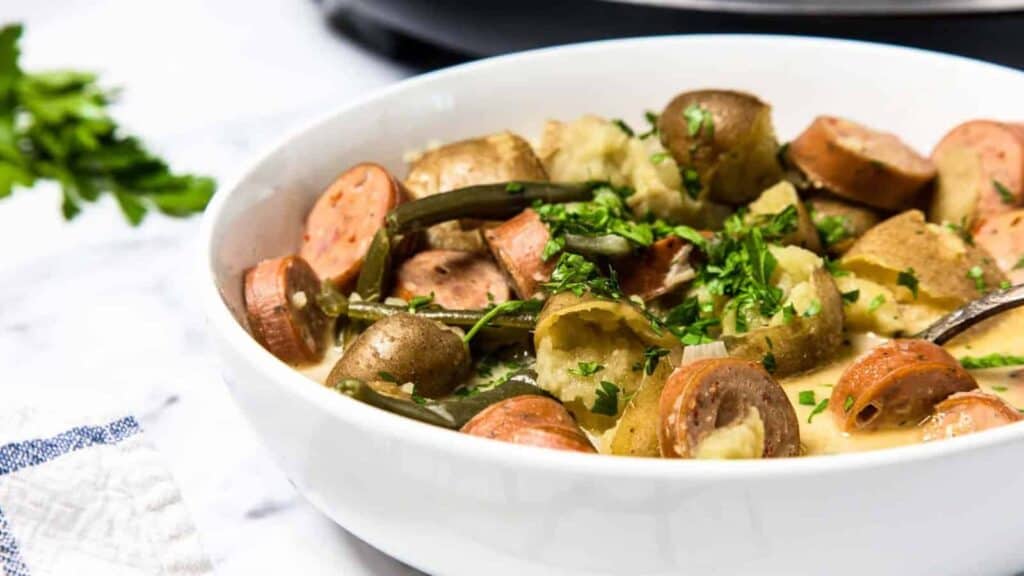 A white bowl filled with sliced sausage, potatoes, and green beans, garnished with chopped parsley.