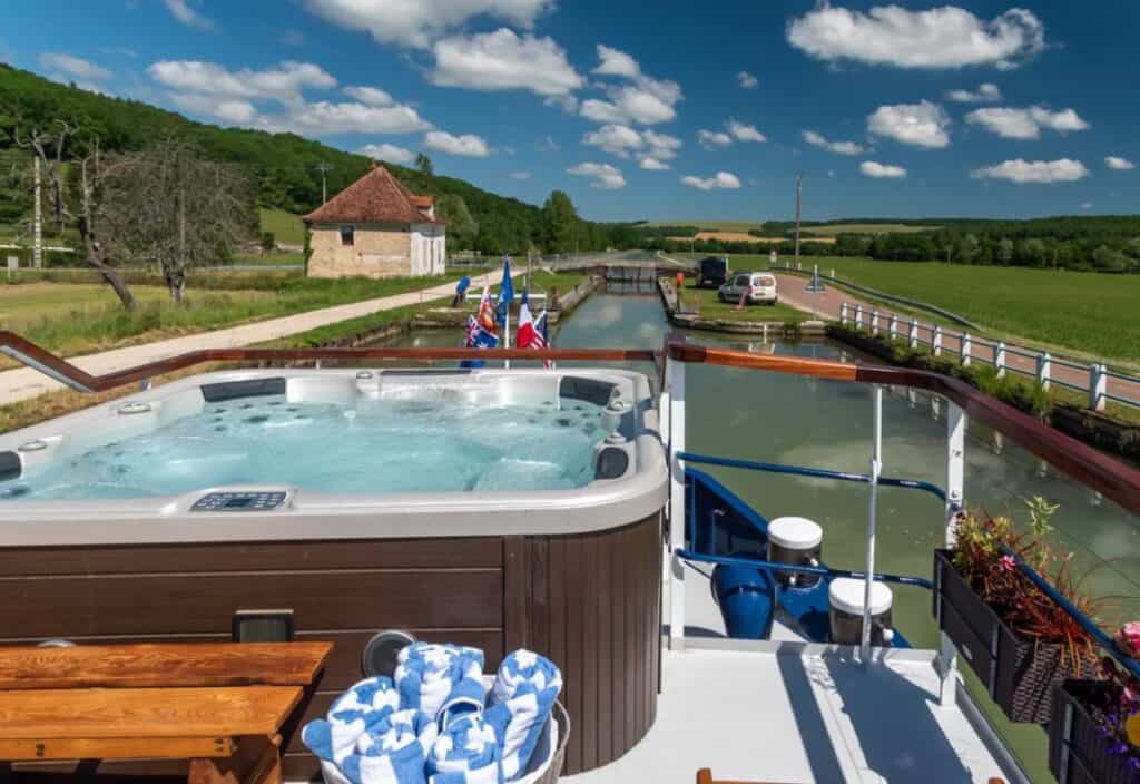 A hot tub on the deck of a luxury barge cruise overlooks a calm waterway, green fields, and a house under a blue sky with scattered clouds.