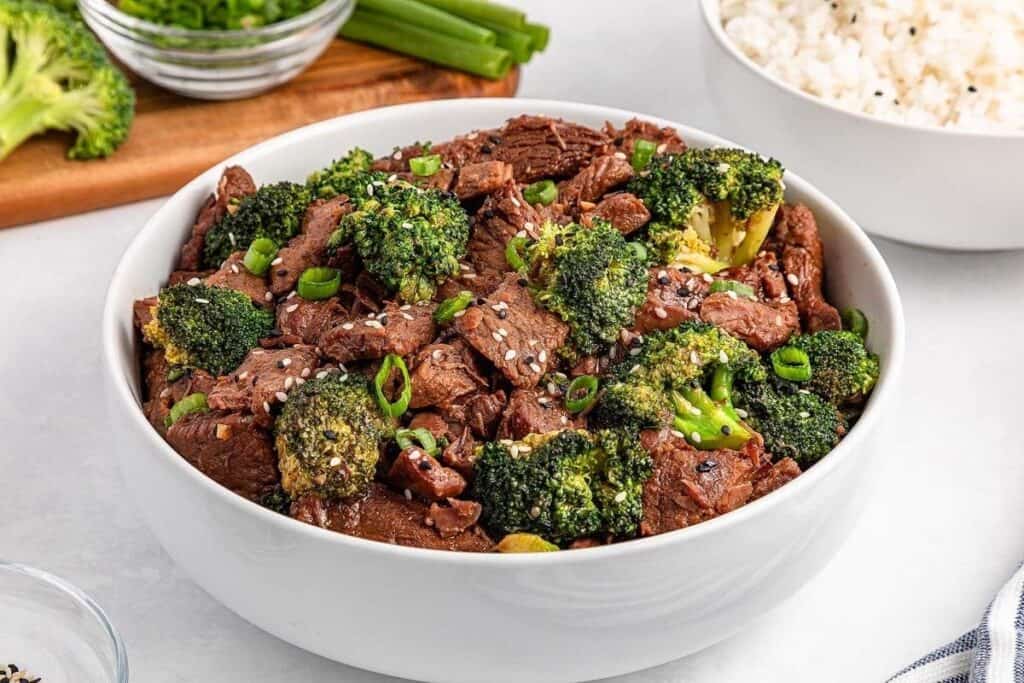A white bowl filled with slow cooked beef and broccoli stir-fry, garnished with sesame seeds and sliced green onions. A bowl of rice and some vegetables are visible in the background.