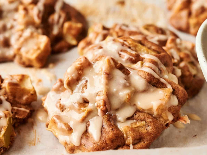 Close-up of freshly baked apple cookies drizzled with icing, resting on parchment paper with part of a bowl visible to the right.