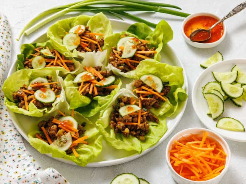 A plate of chicken lettuce wraps filled with shredded carrots and cucumber slices, with bowls of sliced cucumbers, shredded carrots, and dipping sauce on the side.