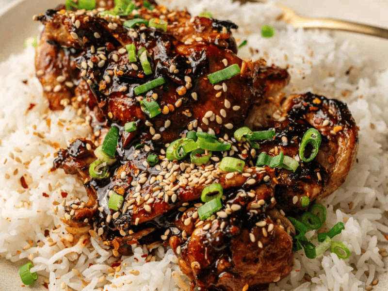 A plate of white rice topped with glazed chicken pieces, garnished with sesame seeds and chopped green onions.