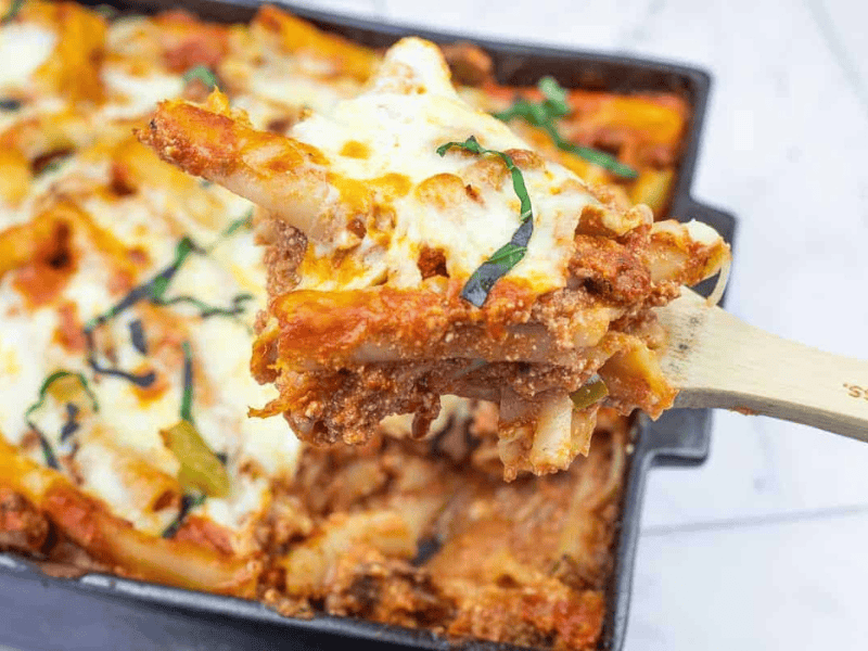 A close-up of a wooden spoon lifting a serving of baked ziti with melted cheese and tomato sauce from a casserole dish.