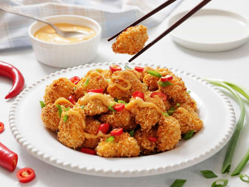 A plate of Bang Bang Chicken Bites garnished with sliced red chili, green onions, and drizzled sauce, with a bowl of dipping sauce in the background.