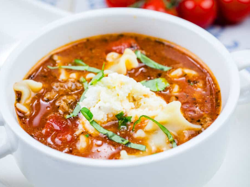 A bowl of beefy lasagna soup with a dollop of ricotta cheese, garnished with basil.