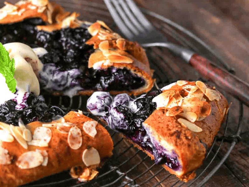 A sliced blueberry galette topped with almond flakes and a scoop of vanilla ice cream, served on a wire cooling rack with a fork nearby.