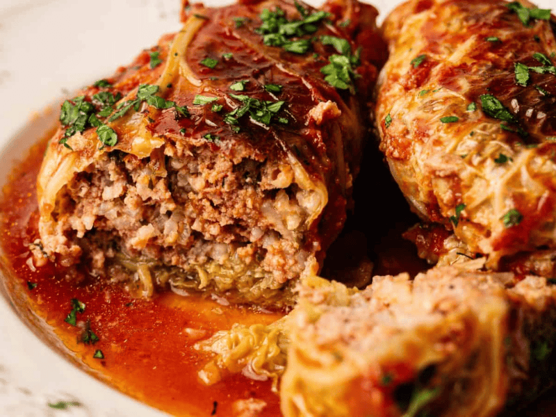 Two cabbage rolls filled with ground meat and rice, topped with tomato sauce and chopped herbs, served on a white plate.
