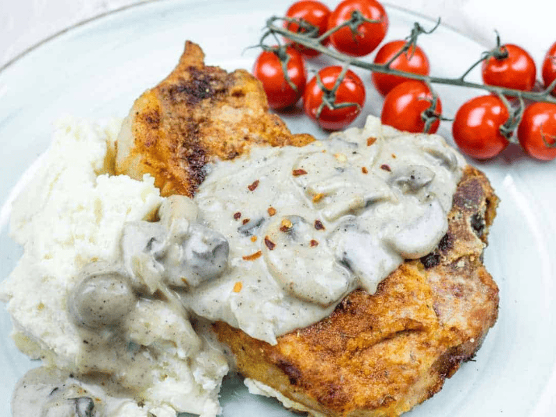 Plate with Cast Iron Smothered Pork Chops served alongside mashed potatoes and vine tomatoes.