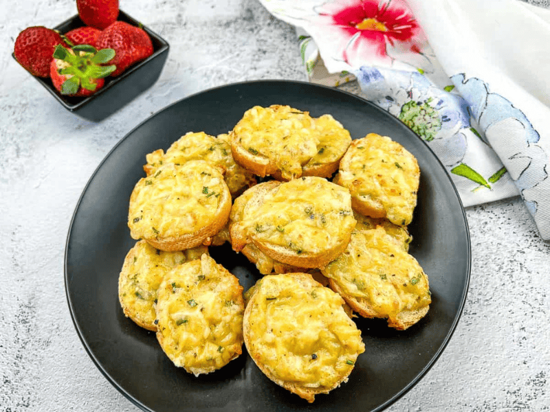 Bite-sized Cheese Toasties on a black plate with a small bowl of strawberries and a floral napkin nearby.