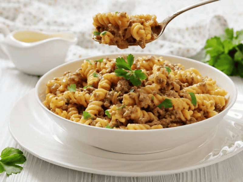 A white bowl of Cheeseburger Hamburger Helper, garnished with parsley, sits on a white plate, with a fork holding a bite above the bowl.