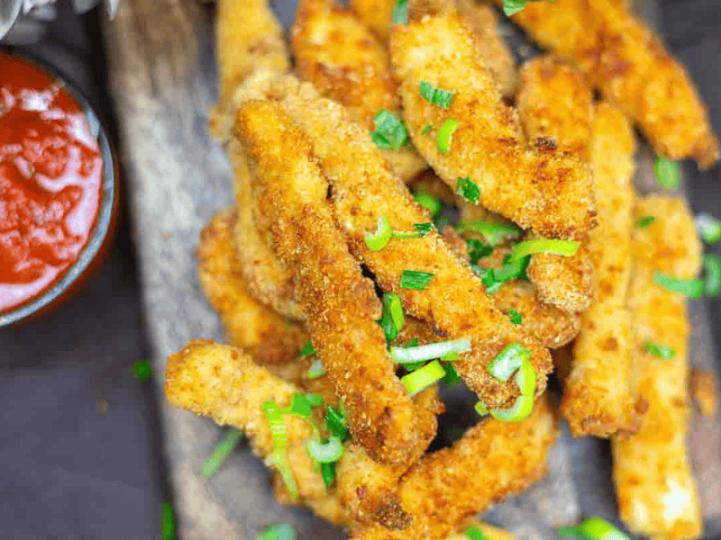 Breaded and fried chicken strips garnished with chopped green onions, served on a wooden board with a side of red dipping sauce.