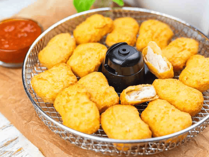 A wire basket filled with golden-brown chicken nuggets, some bitten, with a small cup of red dipping sauce beside it.