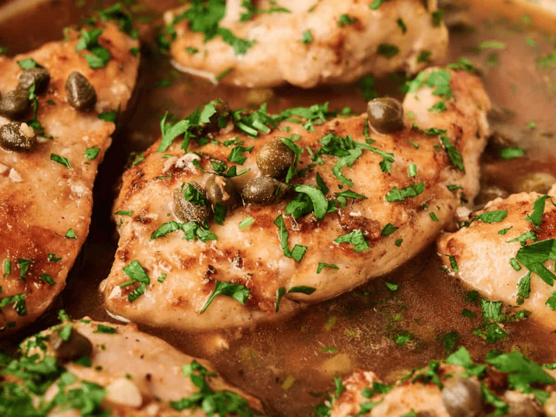 Chicken breasts simmering in a brown sauce, topped with capers and chopped fresh herbs.