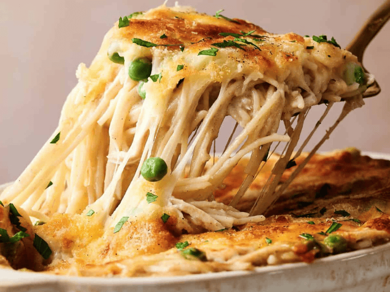 A fork lifts a cheesy serving of baked pasta with peas and shredded chicken from a casserole dish, topped with melted cheese and herbs.
