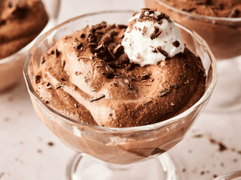 A glass dessert cup filled with chocolate mousse, topped with whipped cream and chocolate shavings, placed on a light-colored surface.