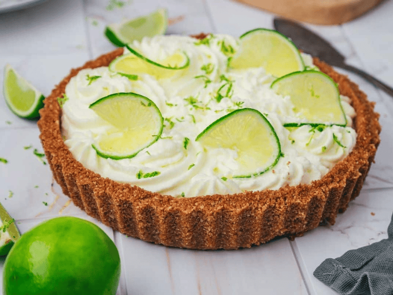 A key lime pie with whipped cream topping, garnished with lime slices and zest, sits on a white surface with additional lime wedges and a knife nearby.