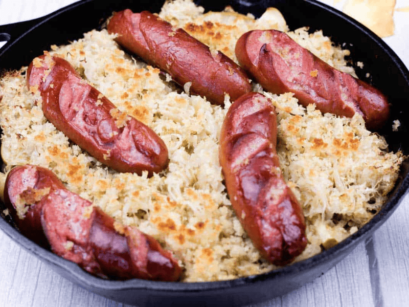 A cast iron skillet filled with sauerkraut and topped with six grilled sausages, all placed on a light wooden surface.