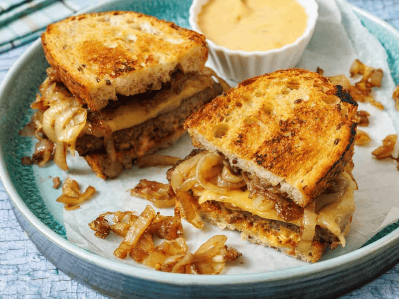 Two halves of a grilled sandwich with melted cheese, caramelized onions, and meat on toasted bread, served on a plate with a side of dipping sauce.