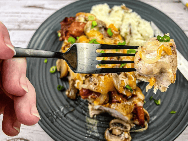 A hand holds a fork with a bite of cheesy baked chicken and vegetables above a plate of the same dish, garnished with green onions and served with rice.