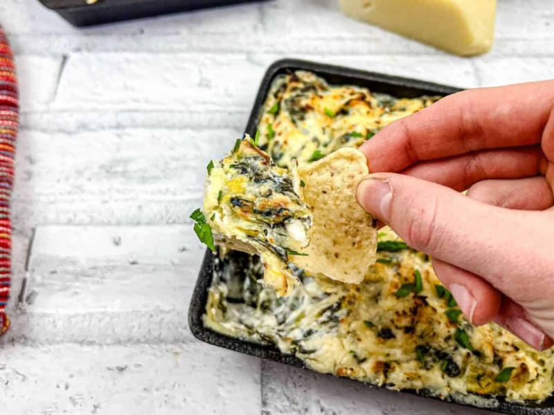 A hand dipping a tortilla chip into a tray of Copycat Applebee&rsquo;s spinach artichoke dip.