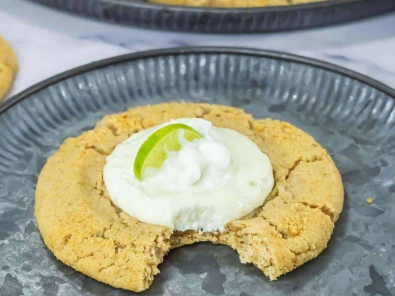 A large Copycat Crumbl Key Lime Cookies with a bite taken out, topped with white cream and a lime wedge, served on a gray plate.